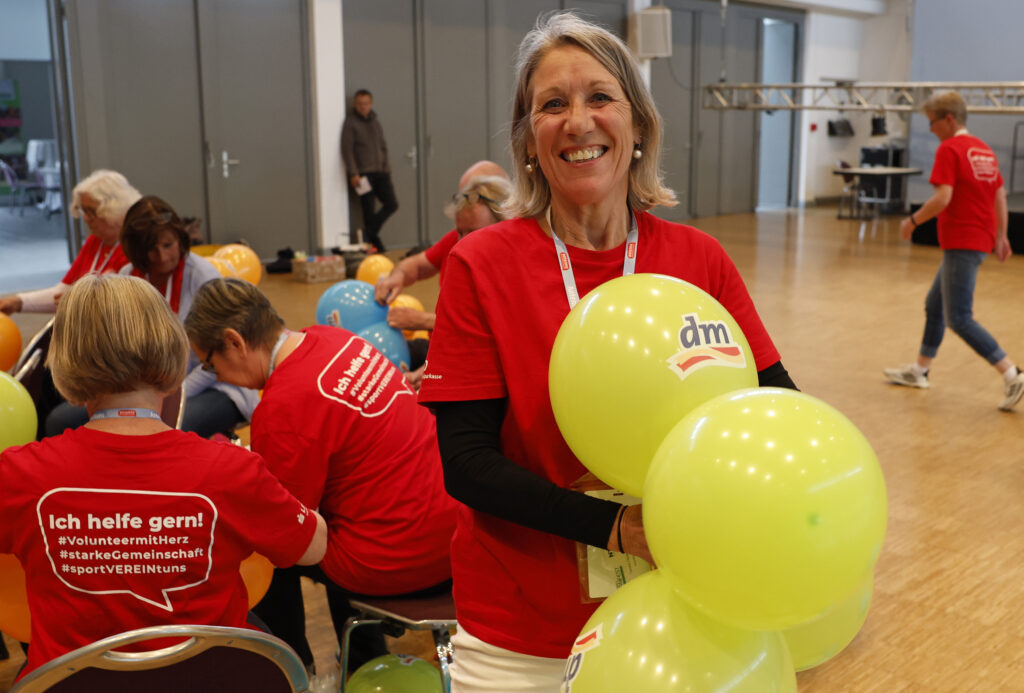 Frau mit einem roten T-Shirt, die lächelt und gelbe Luftballons in der Hand hält. Im Hintergrund sieht man weitere Frauen, die Luftballons knoten.
