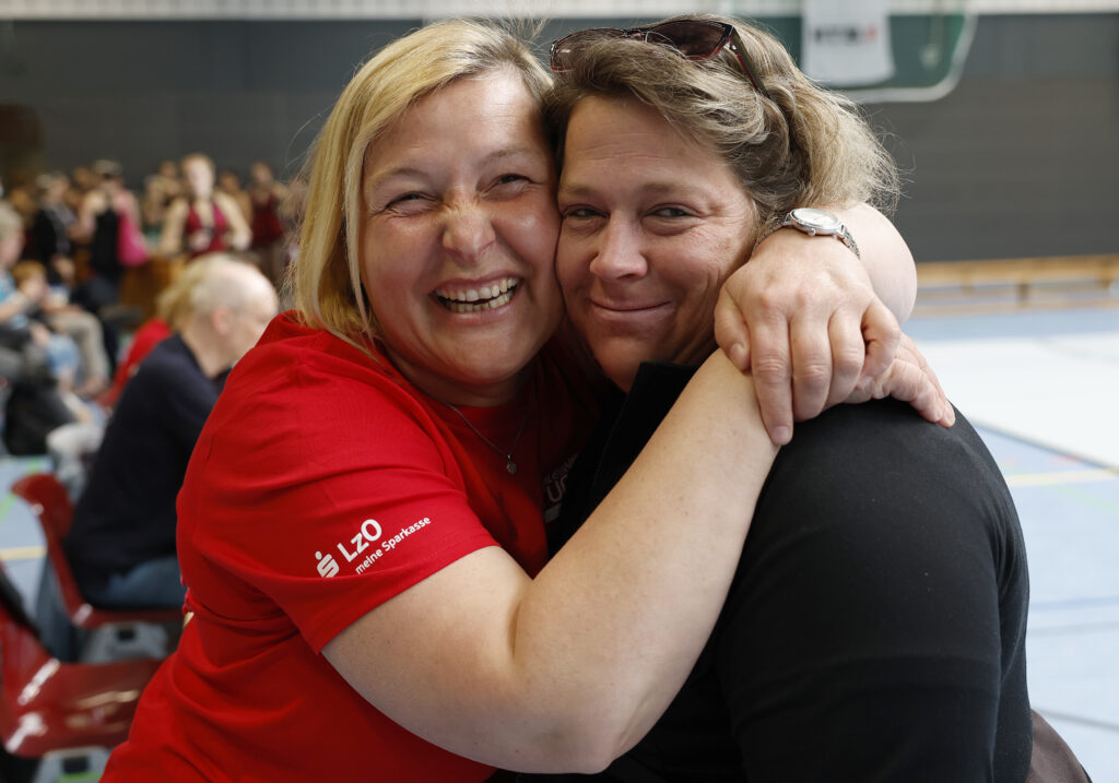 Zwei Frauen umarmen sich und schauen dabei lächelnd in die Kamera.