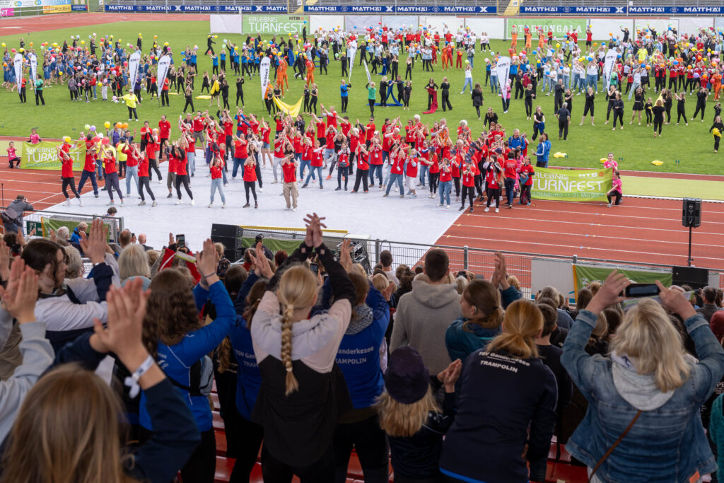 Es ist ein Teil einer Rasenfläche eines Stadios zu sehen. Darauf stehem viele Personen in einem roten T-Shirt und winken dem Publikum zu.