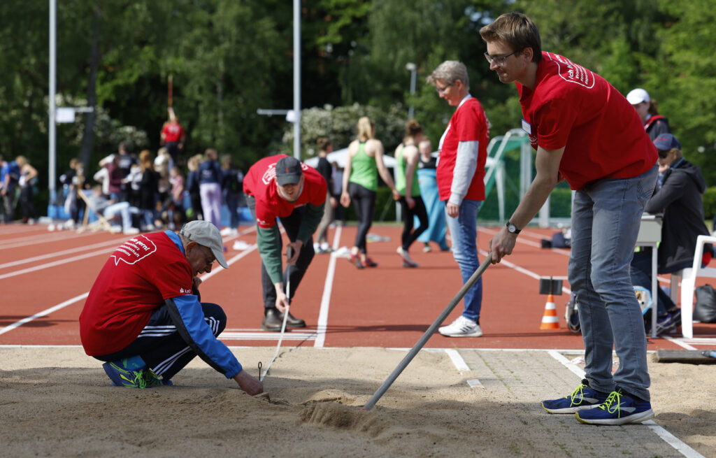 Drei Personen glätten den Stand in einer Weitsprunggrube. Dabei Tragen Sie rote Tshirts.