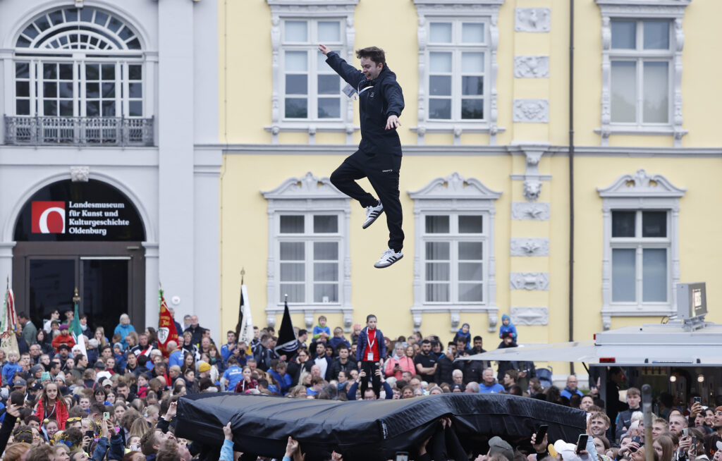 Ein junger Mann springt bei einer Outdoor-Aktion in ein gespanntes Luftkissen, umringt von einer großen Menschenmenge.