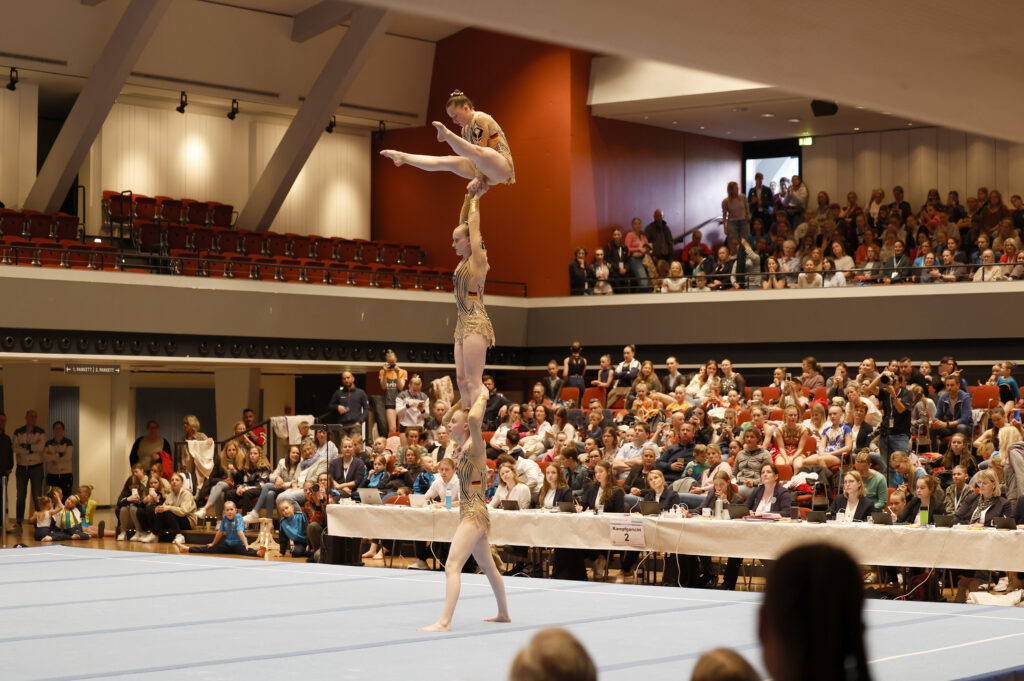 Drei Turnerinnen zeigen eine anspruchsvolle Hebefigur vor vollen Zuschauerrängen.