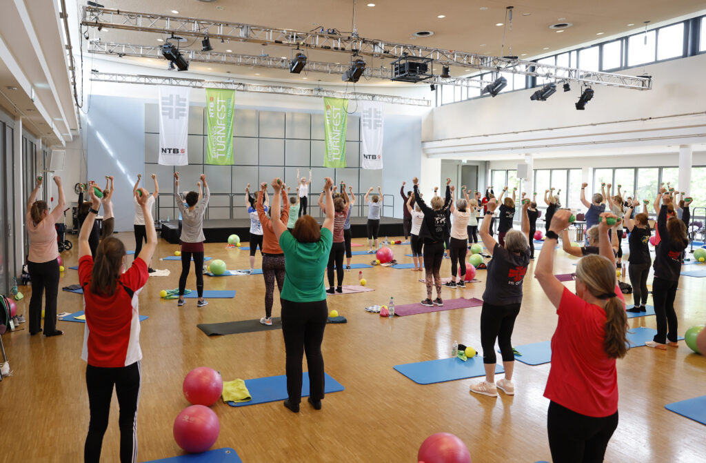 Teilnehmende trainieren gemeinsam mit Hanteln in einer großen Sporthalle.