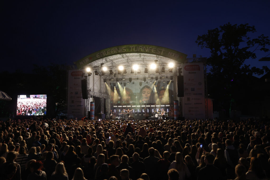 Große Open-Air-Bühne mit Lichtshow und jubelndem Publikum in der Dämmerung.