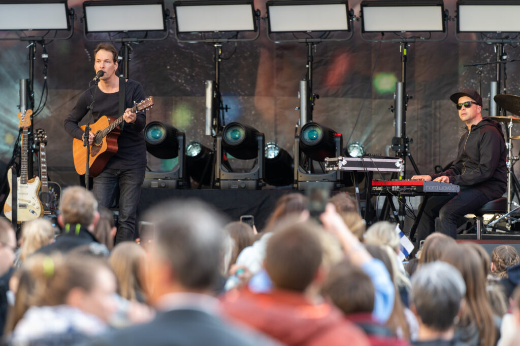 Musiker mit Akustikgitarre und Keyboarder treten vor einem großen Publikum auf einer Open-Air-Bühne auf.