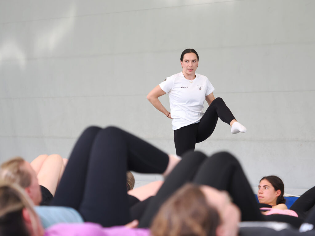Trainerin leitet eine Dehnübung vor liegenden Teilnehmerinnen in der Halle an