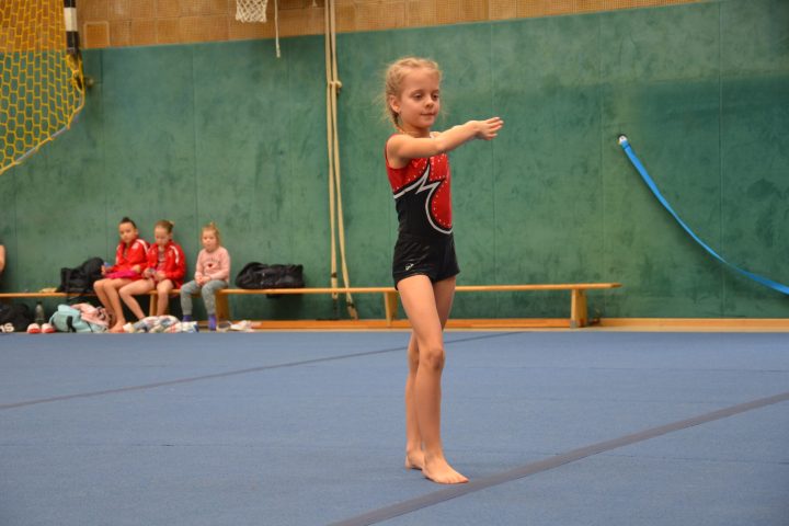 Junges Mädchen in einem Turnanzug führt eine Übung auf einer blauen Bodenmatte in einer Sporthalle vor, während andere Kinder im Hintergrund auf einer Bank sitzen und zuschauen.