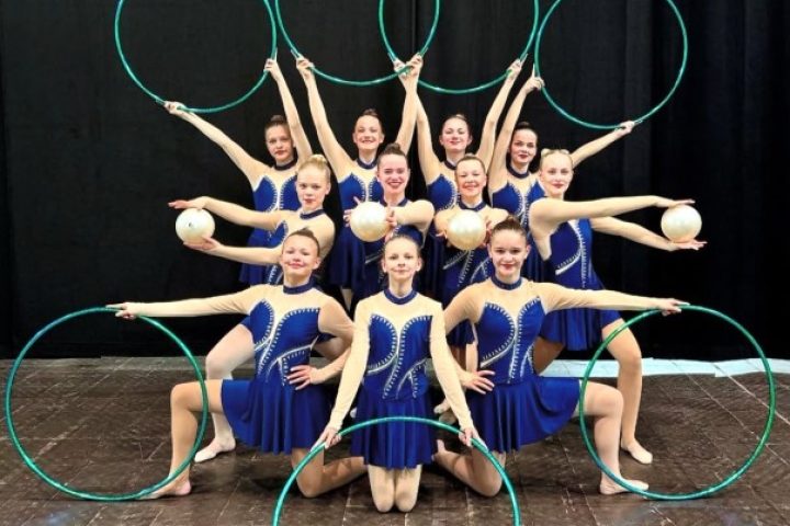 Eine Gruppe von Tänzerin in einem blauen Anzug die mit Bällen und Reifen lächeln.