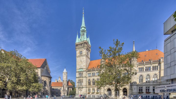 Das Rathaus mit seinem hohen Turm und grünen Turmspitze an einem sonnigen Tag; im Vordergrund ein Wasserspiel auf dem Platz vor dem Rathaus, links und rechts davon grüne Laubbäume. Links im Hintergrund ist der Turm der Burg Dankwarderode zu erkennen.