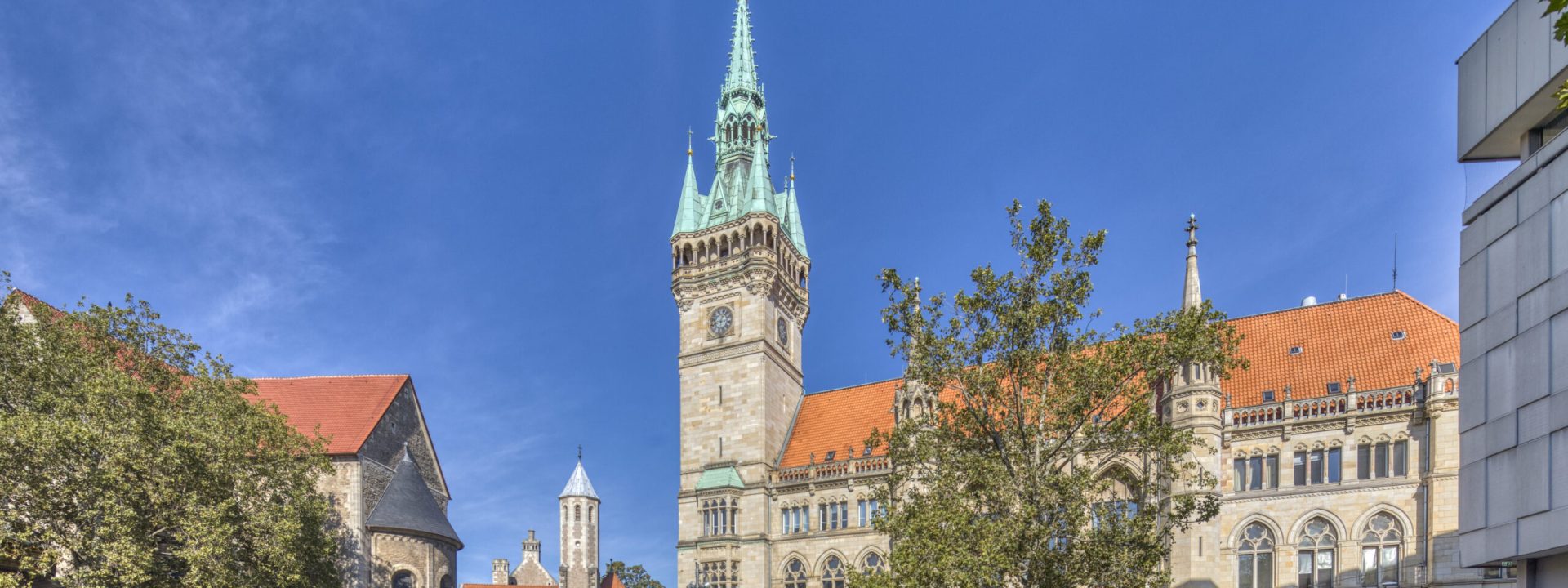 Das Rathaus mit seinem hohen Turm und grünen Turmspitze an einem sonnigen Tag; im Vordergrund ein Wasserspiel auf dem Platz vor dem Rathaus, links und rechts davon grüne Laubbäume. Links im Hintergrund ist der Turm der Burg Dankwarderode zu erkennen.