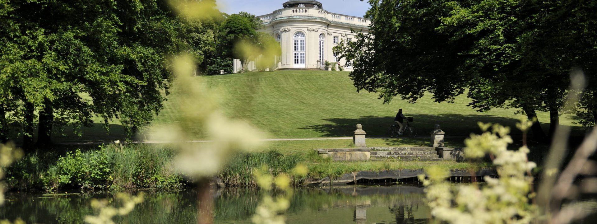 Das helle Schloss Richmond auf einer grünen Anhöhe, davor ein ruhiger Teich, in dem sich das Gebäude spiegelt, eingerahmt von Bäumen und Sträuchern.