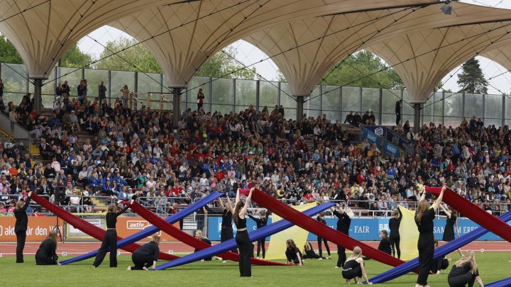 Eine große Tribüne mit vielen Zuschauern im Hintergrund. Im Vordergrund sieht man Personen, die mit Tüchern ein Bild erzeugen
