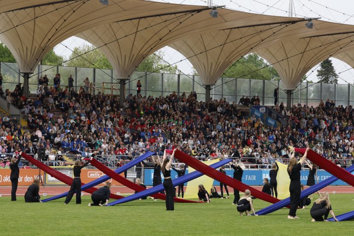 Eine große Tribüne mit vielen Zuschauern im Hintergrund. Im Vordergrund sieht man Personen, die mit Tüchern ein Bild erzeugen