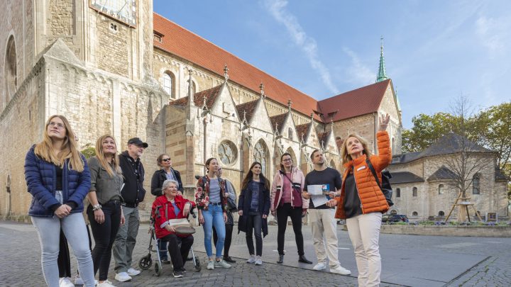 - Eine Gästeführerin in oranger Jacke zeigt und erzählt einer Gruppe Besucher:innen am Dom St. Blasii etwas ; die Teilnehmenden – darunter eine Person mit Rollator – schauen aufmerksam in dieselbe Richtung.