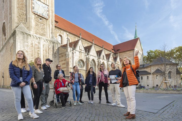 - Eine Gästeführerin in oranger Jacke zeigt und erzählt einer Gruppe Besucher:innen am Dom St. Blasii etwas ; die Teilnehmenden – darunter eine Person mit Rollator – schauen aufmerksam in dieselbe Richtung.