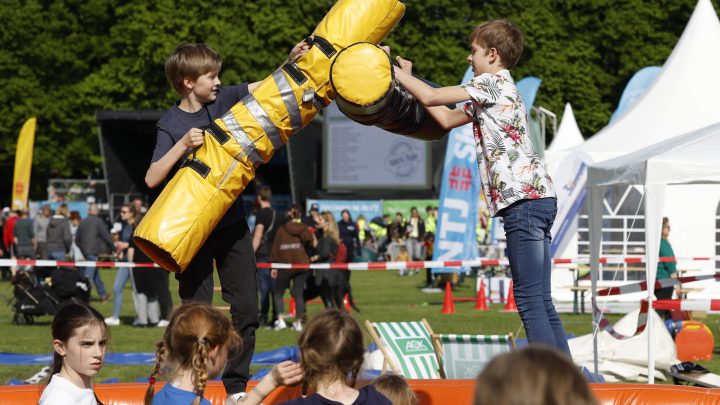 Zwei Jungen kämpfen spielerisch mit gepolsterten Stäben auf einer Eventfläche, während andere Kinder zuschauen.