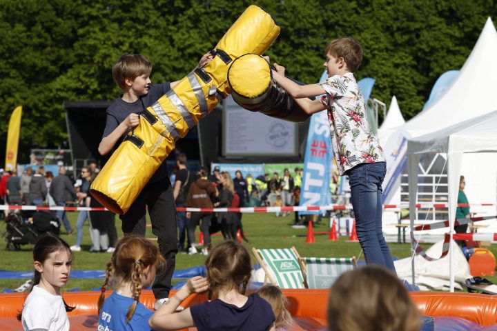 Zwei Jungen kämpfen spielerisch mit gepolsterten Stäben auf einer Eventfläche, während andere Kinder zuschauen.