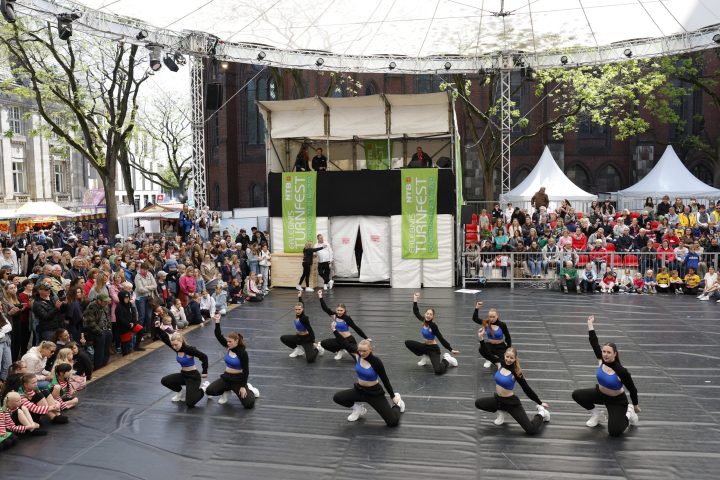 Eine Gruppe von Tänzern in gleichem Outfit stehen auf einer runden Bühne