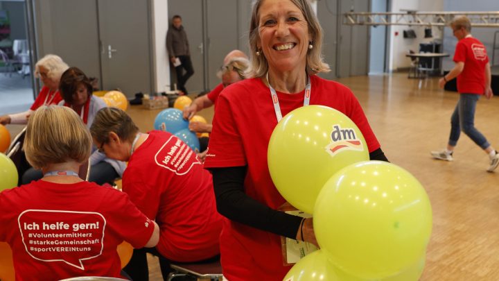 Frau mit einem roten T-Shirt, die lächelt und gelbe Luftballons in der Hand hält. Im Hintergrund sieht man weitere Frauen, die Luftballons knoten.