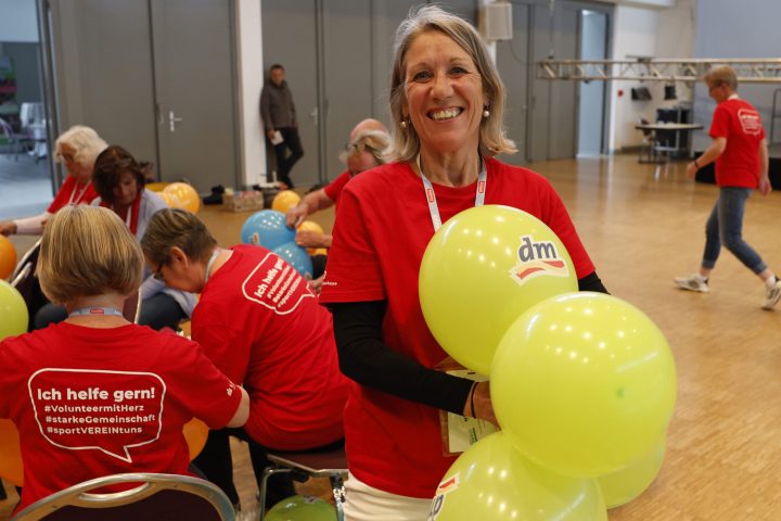 Frau mit einem roten T-Shirt, die lächelt und gelbe Luftballons in der Hand hält. Im Hintergrund sieht man weitere Frauen, die Luftballons knoten.