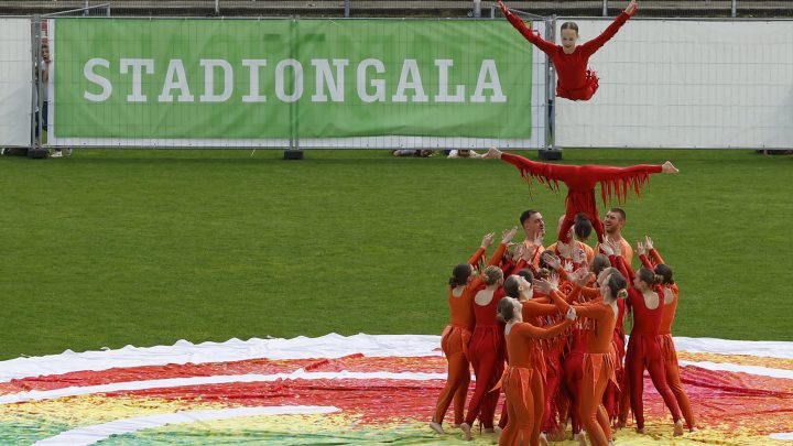 Akrobatikgruppe in roten Kostümen präsentiert eine spektakuläre Hebefigur auf dem Rasen während der Stadiongala.