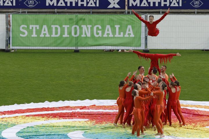 Akrobatikgruppe in roten Kostümen präsentiert eine spektakuläre Hebefigur auf dem Rasen während der Stadiongala.