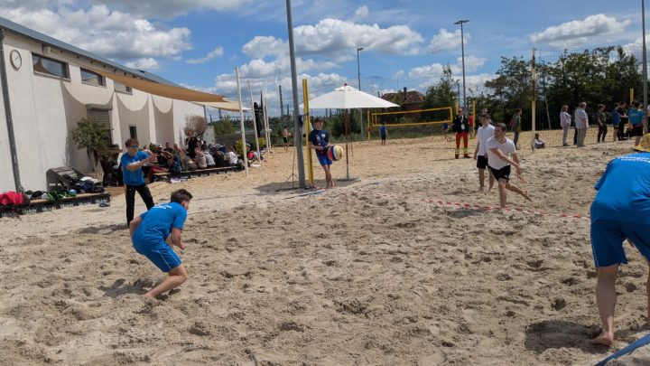 Teams spielen Beach-Völkerball auf einem Sandfeld unter freiem Himmel.