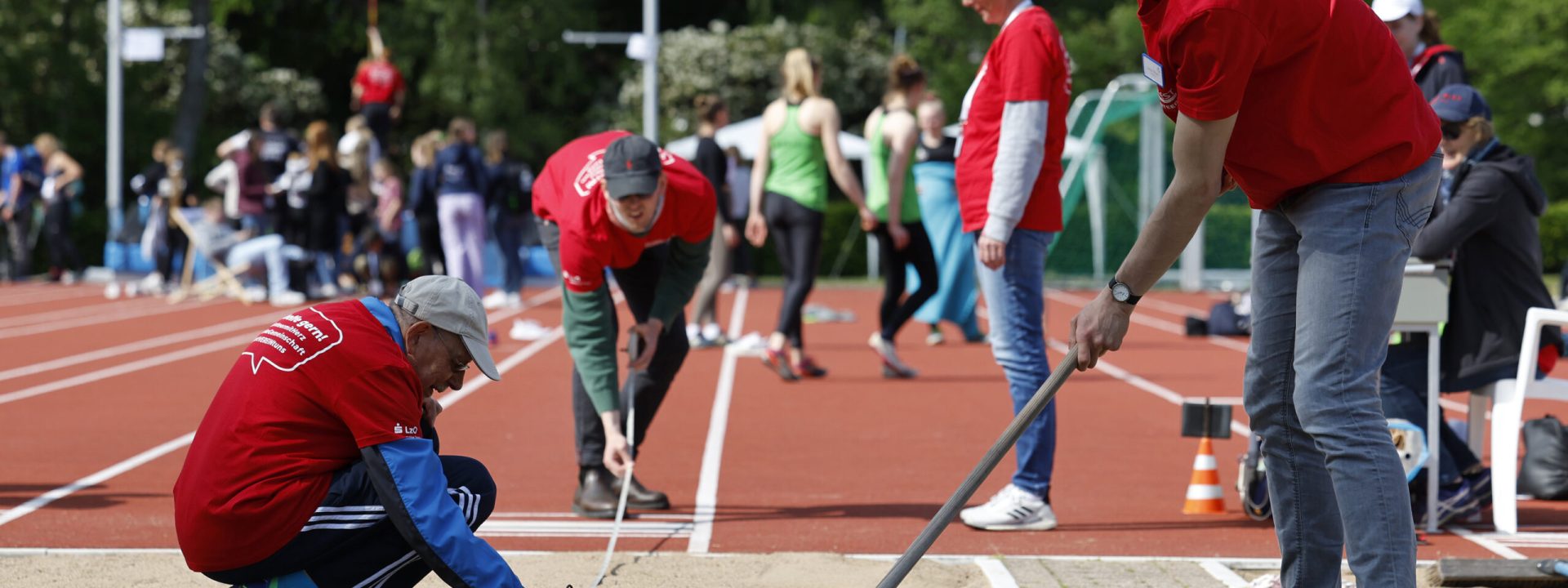 Drei Personen glätten den Stand in einer Weitsprunggrube. Dabei Tragen Sie rote Tshirts.
