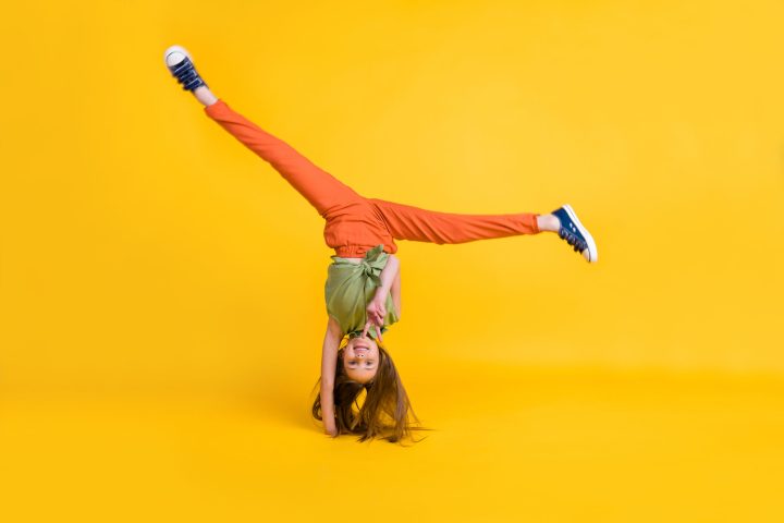 Full length body size photo little girl wearing casual clothes careless doing handstand isolated vivid yellow color background
